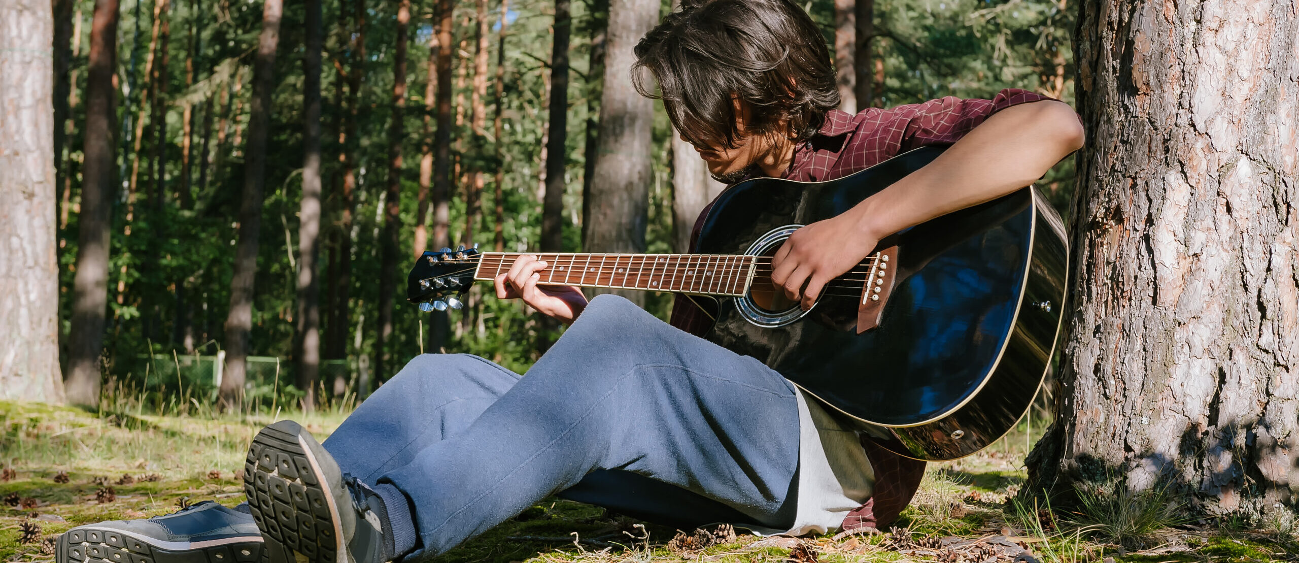 A man plays the guitar while sitting in the forest near a tree. Against the backdrop of nature.